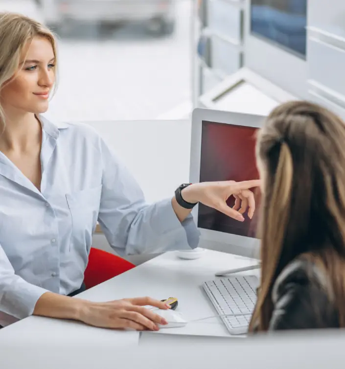 main image webp A woman in a light blue shirt gestures towards a computer screen while speaking to another seated individual.